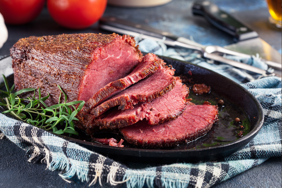 Sliced beef sirloin tip roast on a black oval platter with garnish of green herbs, set on a blue-and-white plaid napkin with knives and tomatoes in the background.