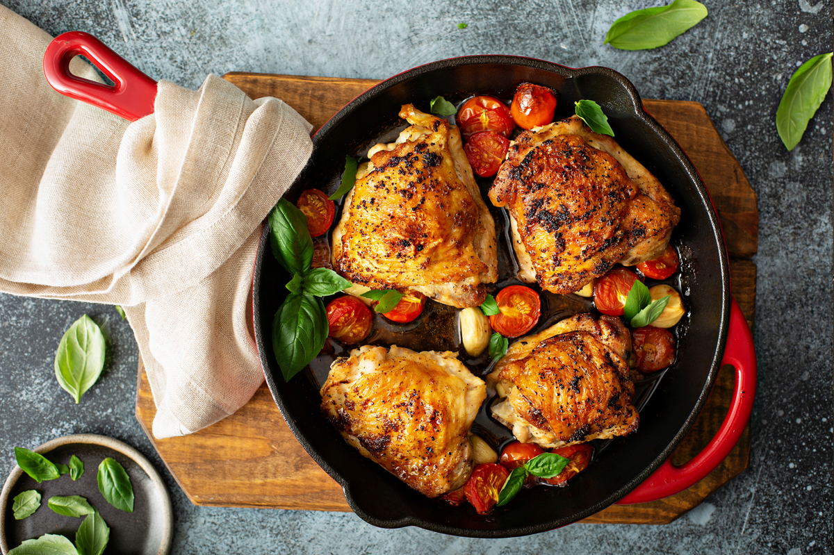 Overhead view of four roasted chicken thighs in a black cast iron skillet with red handles, cooked with halved cherry tomatoes, garlic, and fresh basil on a wooden board with a beige cloth.