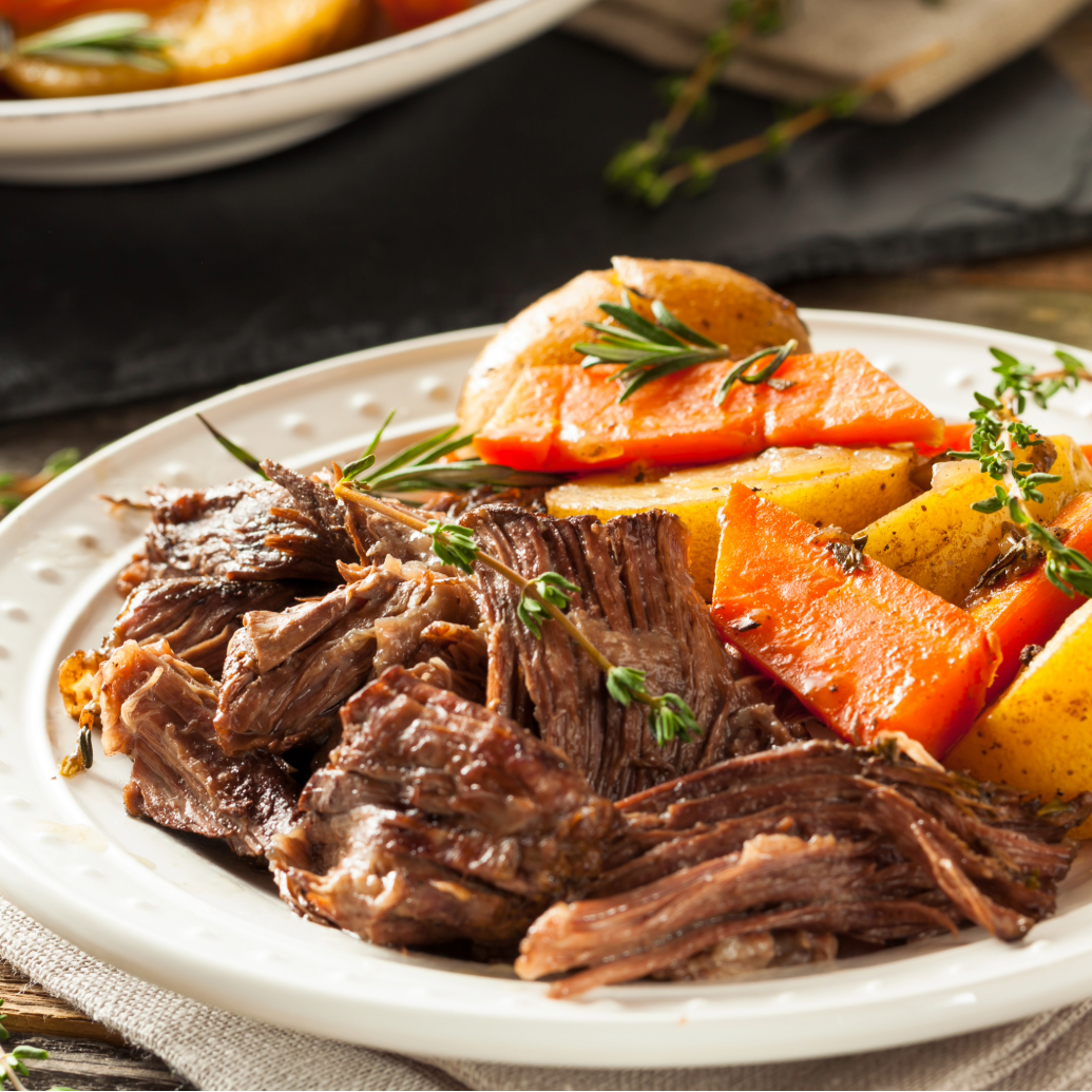 A plated meal of shredded cooked beef with roasted carrots and potatoes, garnished with herbs, set on a wooden table with a fork, salt and pepper shakers, and another serving bowl in the background.