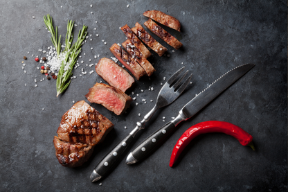 Grilled sirloin steak slices and a seared piece arranged on a dark stone surface with a fork, knife, rosemary, coarse salt, peppercorns, and a red chili pepper.