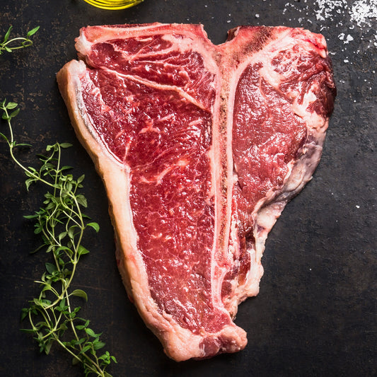 A raw T-bone steak with marbled red meat and white fat, photographed from above on a dark textured surface with a sprig of green herbs and a small amount of oil at the top edge.