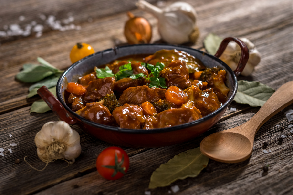 A hearty meat and vegetable stew in a dark cast-iron skillet with wooden handles, set on a rustic wooden table surrounded by garlic, bay leaves, a tomato, and a wooden spoon.