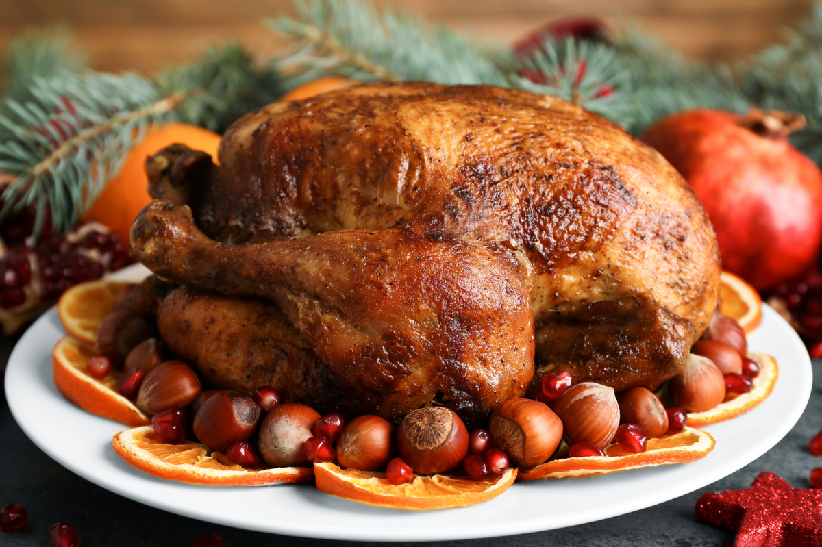 A roasted whole turkey on a white plate, surrounded by orange slices, cranberries, pecans, and pine branches in a festive holiday setting.