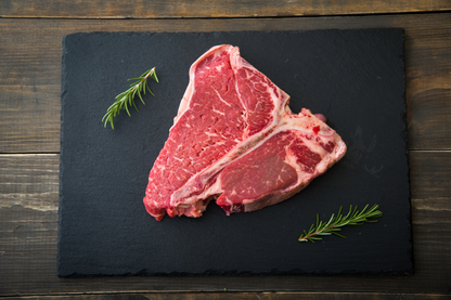 A raw porterhouse steak with visible marbling, placed on a dark rectangular serving board with two sprigs of rosemary, set on a dark wooden surface.