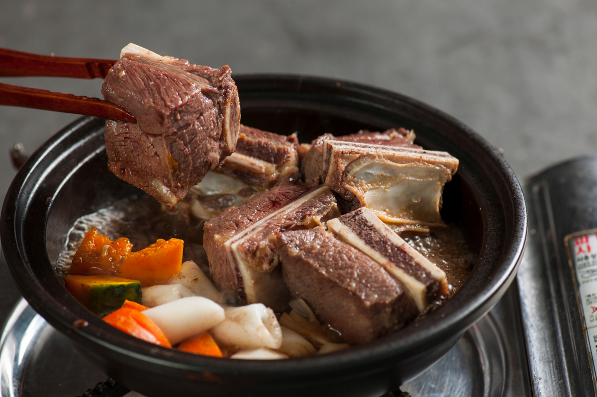 A close-up of a black clay pot on a stove, with chopsticks lifting a piece of braised bone-in meat. The pot contains broth and vegetables, including orange carrots, white radish, and green zucchini, against a gray background.