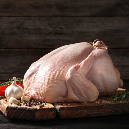 A raw whole turkey placed on a rustic wooden cutting board with garlic, tomatoes, rosemary, and peppercorns, set against a dark wood plank background.