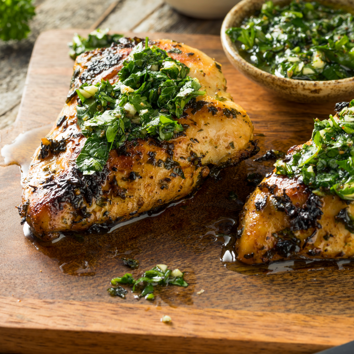 Grilled chicken breast topped with fresh green herb sauce on a wooden cutting board, with a small bowl of the sauce in the background.