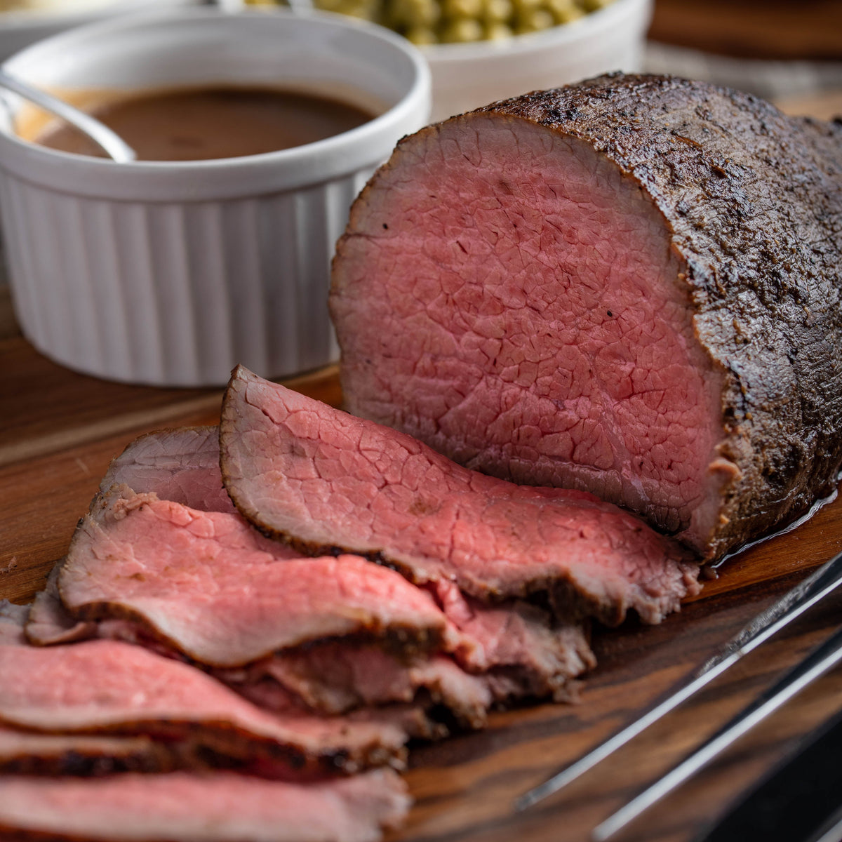 Sliced and whole roasted beef on a wooden serving board, accompanied by bowls of gravy, mashed potatoes, and green peas in a softly blurred dining setting.