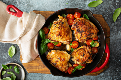 Overhead view of four roasted chicken thighs in a black cast iron skillet with red handles, cooked with halved cherry tomatoes, garlic, and fresh basil on a wooden board with a beige cloth.