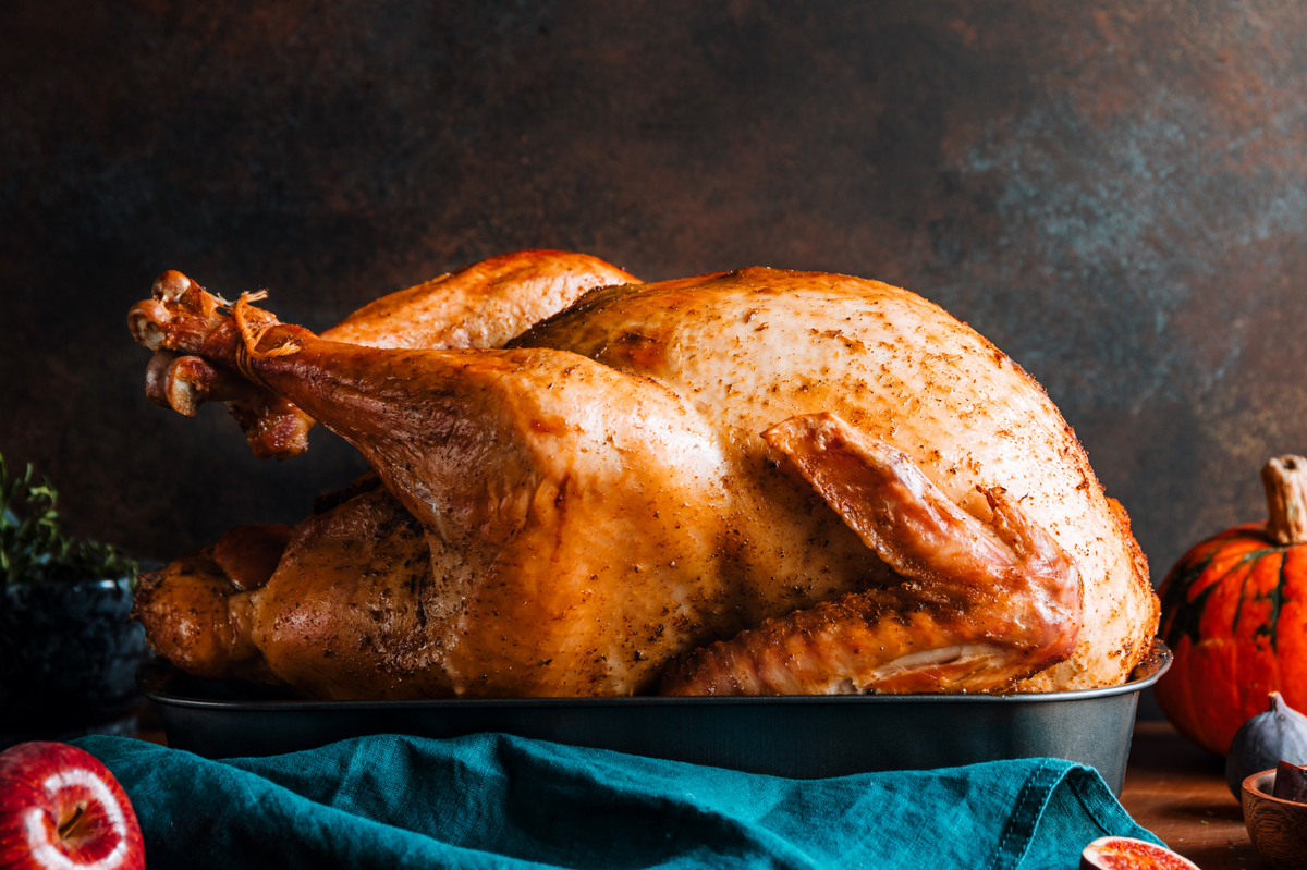 A whole roasted turkey in a dark baking pan, set on a teal cloth with autumn props including a small pumpkin, garlic, and red fruit slices against a dark rustic background.