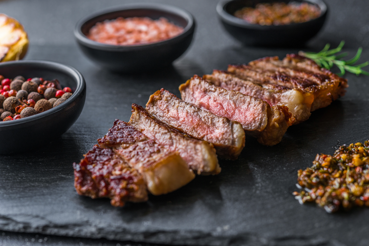 Sliced grilled NY strip steak arranged on a dark slate platter, surrounded by small bowls of spices, a pink condiment, and a herb garnish.