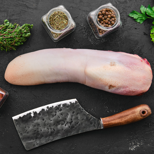 A raw beef tongue presented on a dark textured surface with a cleaver, herbs, and spice jars around it.