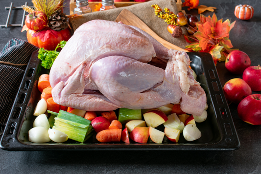 A raw whole turkey placed on a black roasting tray surrounded by chopped apples, carrots, celery, garlic, and autumn decorations on a dark tabletop.