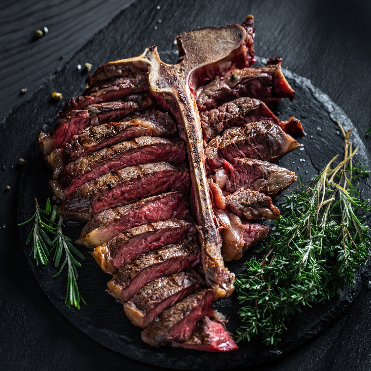 A sliced porterhouse steak served on a dark stone platter with rosemary and thyme garnish, photographed in a dark restaurant-style setting.