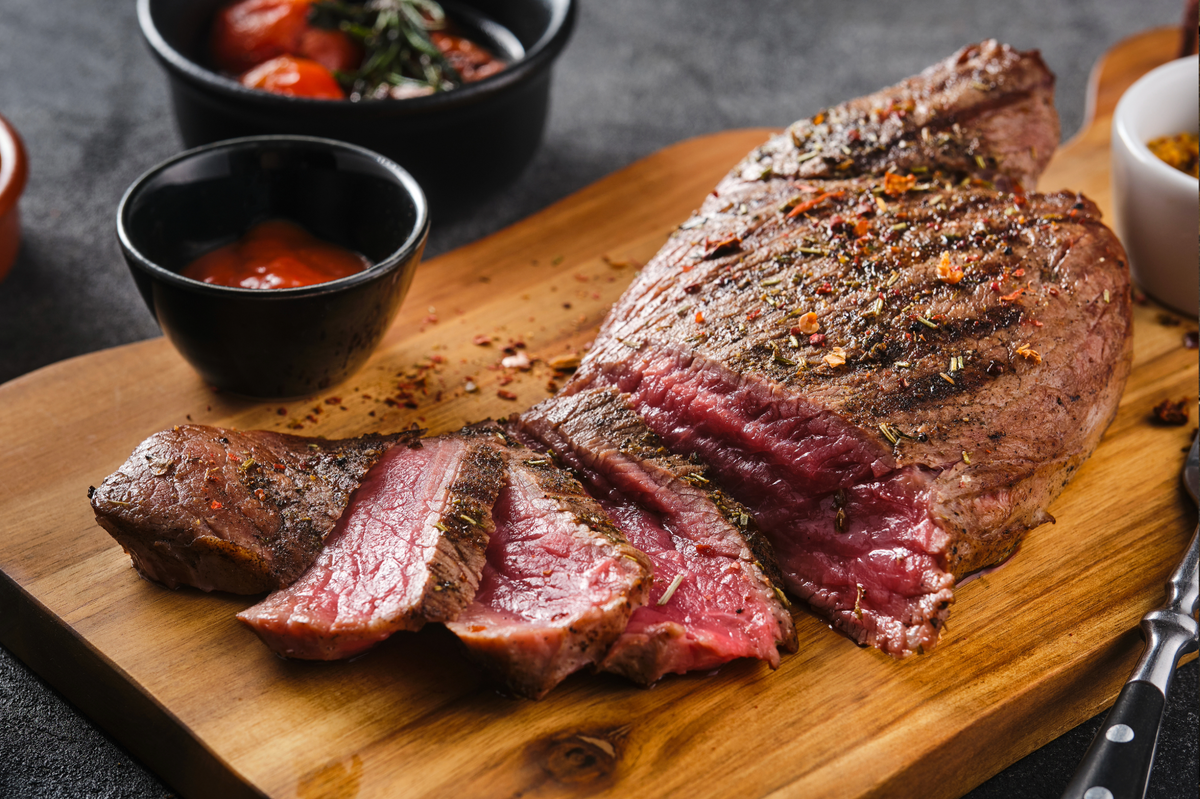 A sliced grilled rump roast with a pink center and charred exterior, served on a wooden cutting board with small bowls of sauce and tomatoes.