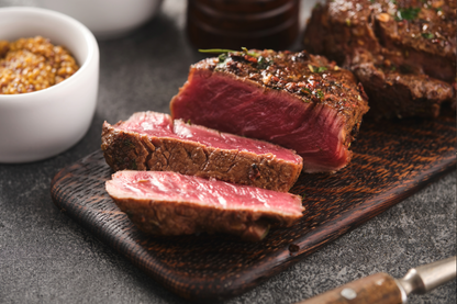 Sliced medium-rare beef filet mignon on a dark wooden cutting board, with a small white bowl of mustard and a dark bottle in the background.