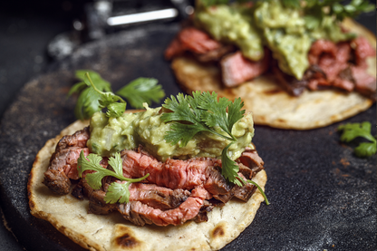 Two prepared tacos on a dark stone plate, topped with sliced cooked flank steak, mashed avocado, and fresh cilantro, photographed in a close-up food setting.