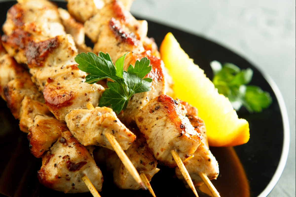 Grilled chicken skewers on wooden sticks served on a black plate with a lemon wedge and parsley garnish.