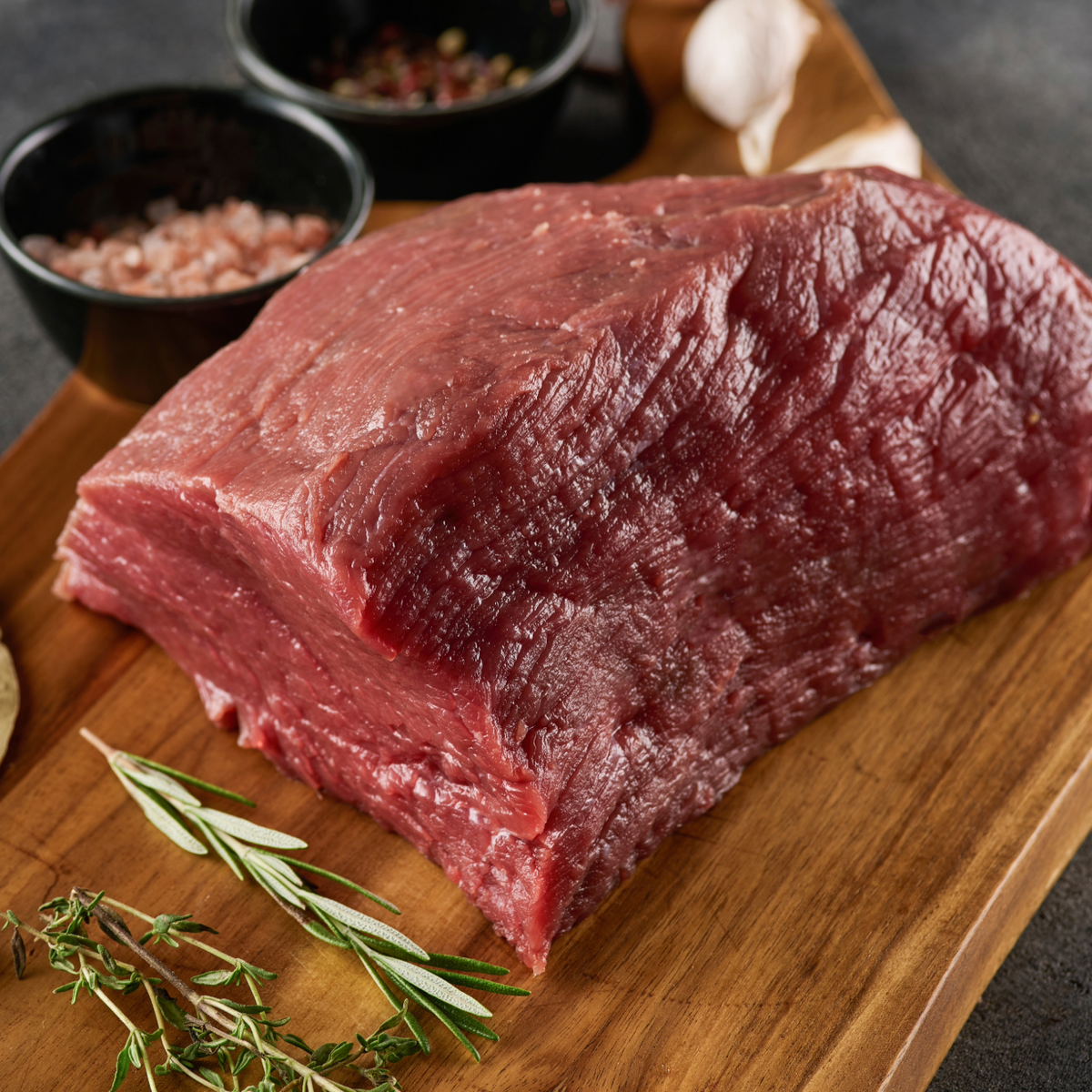 A large raw beef rump roast sits on a wooden cutting board, styled with bay leaves, rosemary, garlic, and small bowls of sea salt and peppercorns on a dark gray surface.