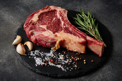 A raw ribeye steak with marbled red meat and fat, served on a black slate platter with garlic cloves, coarse salt, mixed peppercorns, and a sprig of rosemary on a dark textured background.