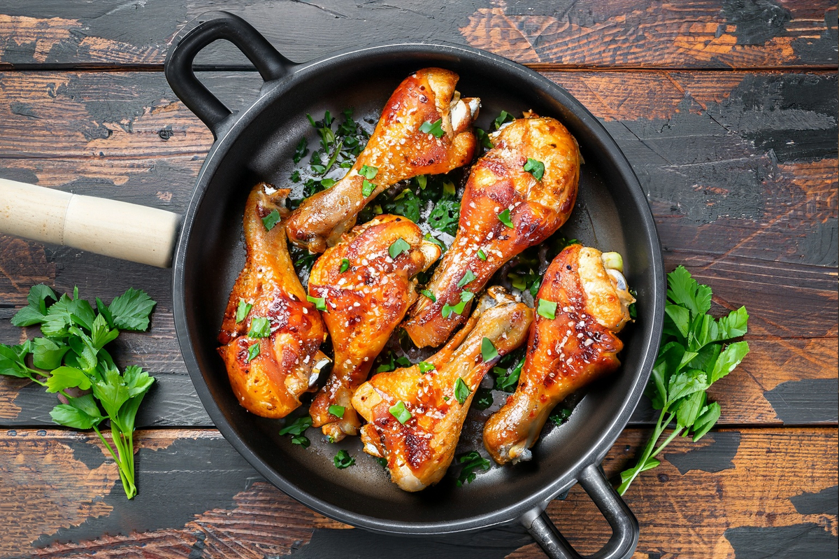 Five cooked chicken drumsticks with a glossy orange-brown glaze and chopped green herbs, presented in a black cast-iron skillet on a rustic dark wooden table with parsley garnish.