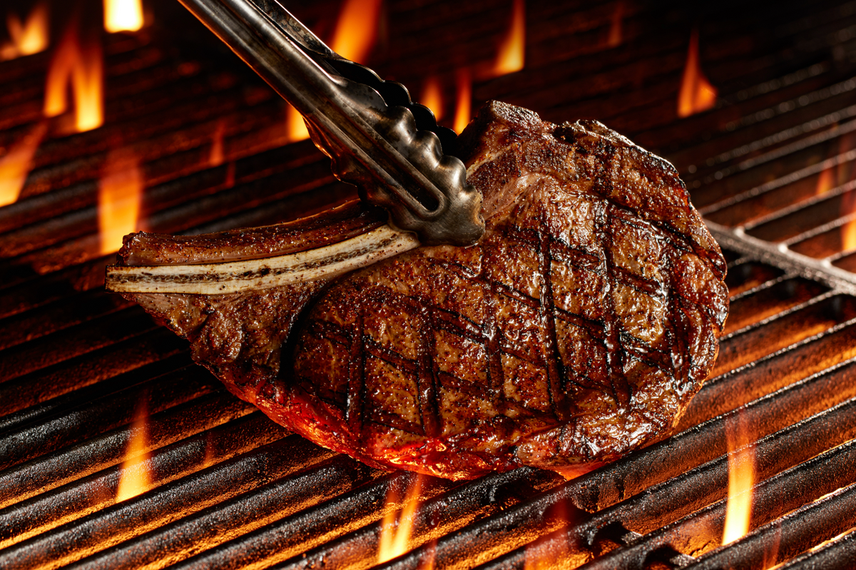A grilled ribeye with a bone and visible flame grill marks, held by metal tongs on a hot barbecue grill.