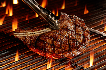 A grilled ribeye with a bone and visible flame grill marks, held by metal tongs on a hot barbecue grill.