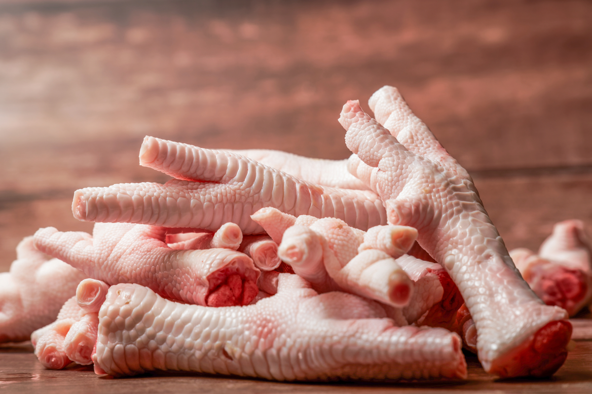 A pile of raw chicken feet with visible skin and joints, arranged on a wooden surface against a warm brown wood background.