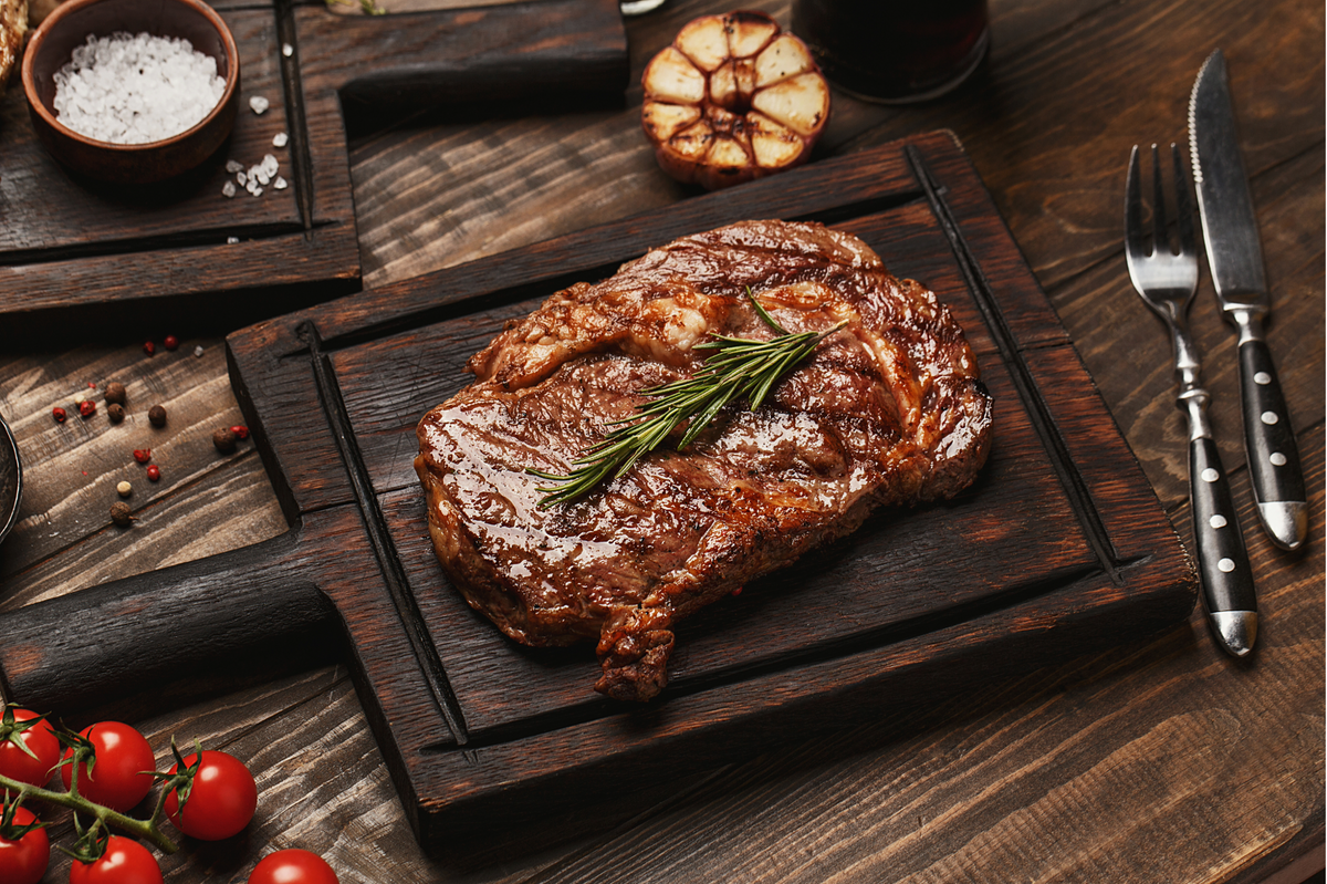 A cooked ribeye steak on a dark wooden cutting board with a sprig of rosemary, styled on a rustic wooden table with a fork, knife, salt, garlic, and cherry tomatoes.