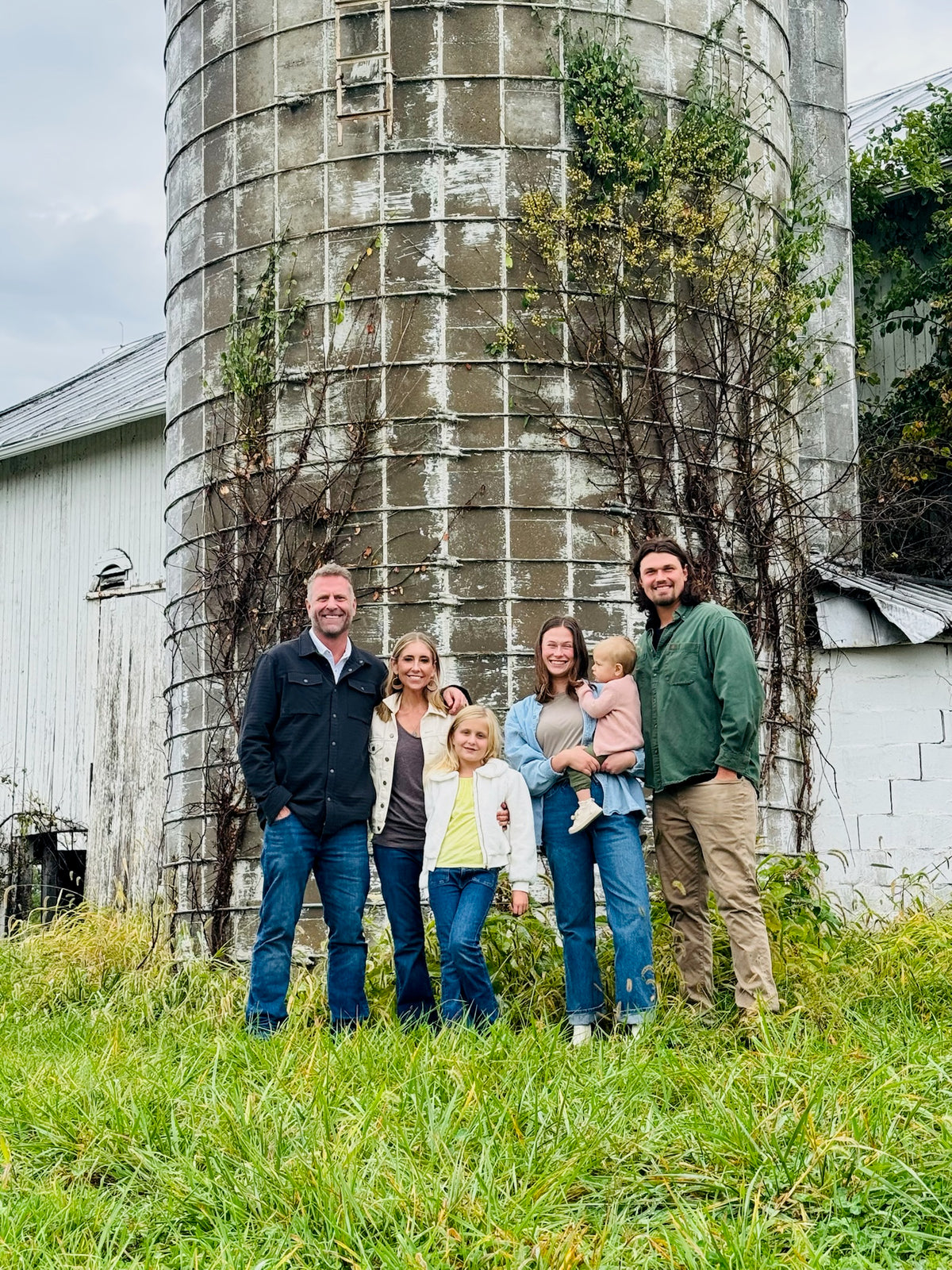 Two families stand together in front of a large weathered silo on a farm, with grass and white buildings in the background.