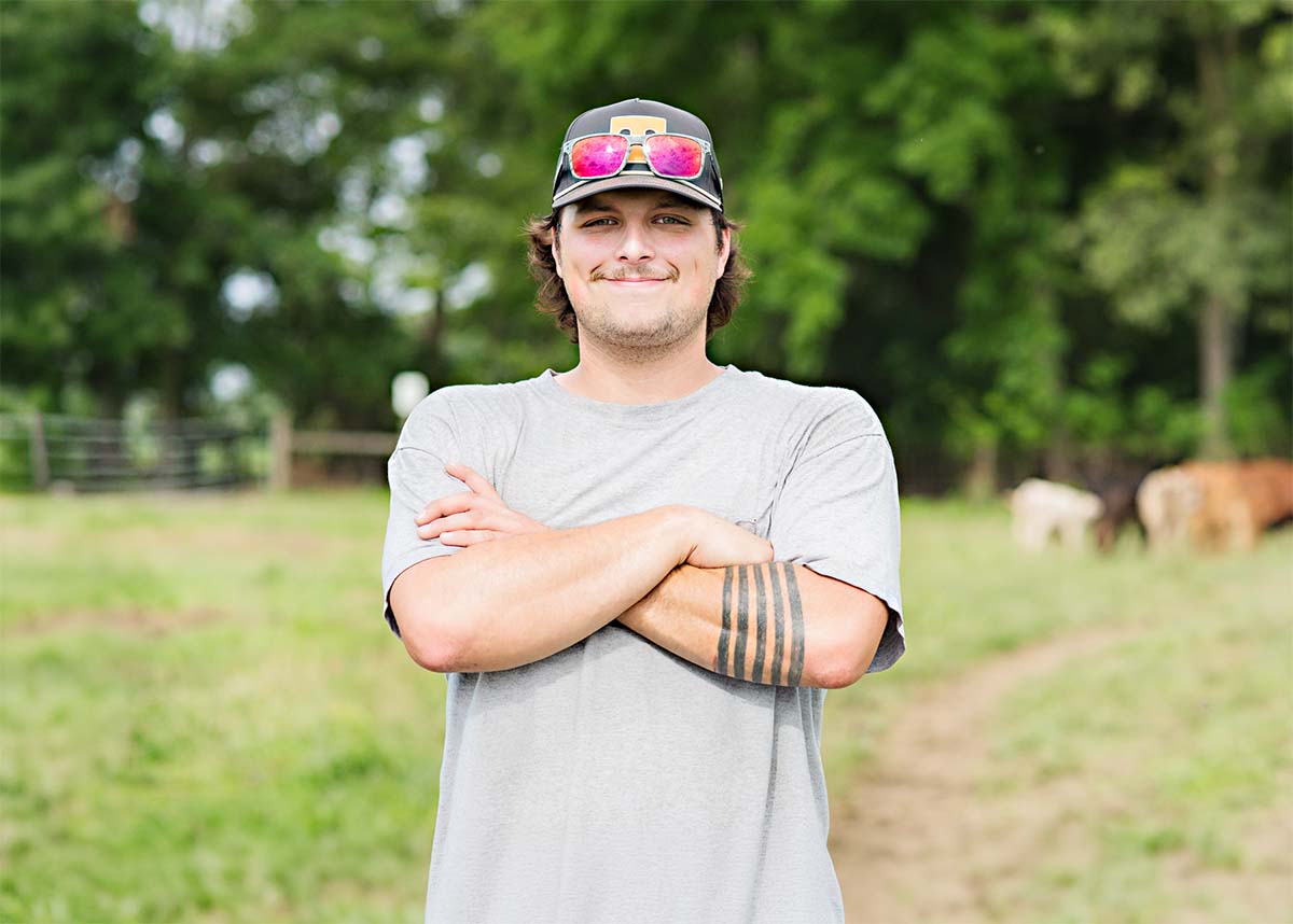 Jason Rutschilling standing in a grassy field with trees in the background, wearing a gray t-shirt, a black baseball cap with pink mirrored sunglasses on the brim, and a black armband tattoo. Two cows are visible in the background on a dirt path.
