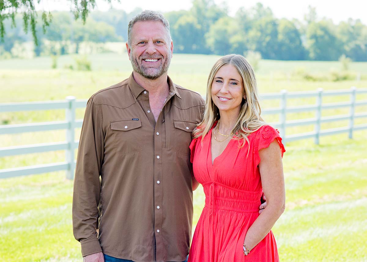 A smiling Charles DeLacey in a brown button-down shirt and a smiling Kelly DeLacey in a bright red dress standing together outdoors in a grassy field with a white fence behind them. Both are co-owners of Yellow Springs Provisions.