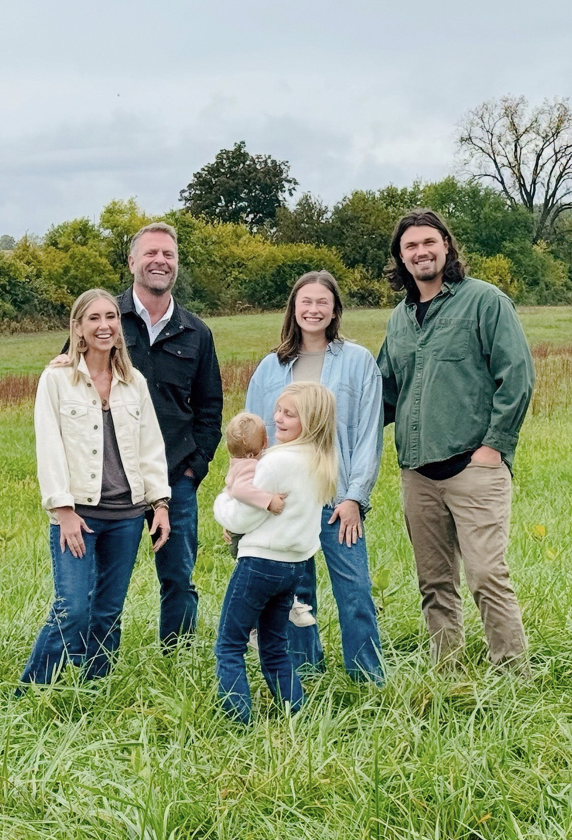 Two families stand together in front of a large open pasture on a farm, with grass and trees in the background.