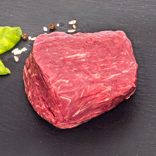 A raw cut of red beef with visible marbling, photographed on a dark slate surface with a few scattered peppercorns and a green herb leaf at the left edge.