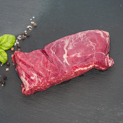 A raw cut of red beef with visible marbling, placed on a dark slate surface with a few green basil leaves and coarse salt and pepper scattered nearby.