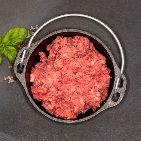 A top-down view of a small round cast iron pot with a metal handle, filled with raw ground meat, set on a dark slate surface with a green herb sprig in the corner.
