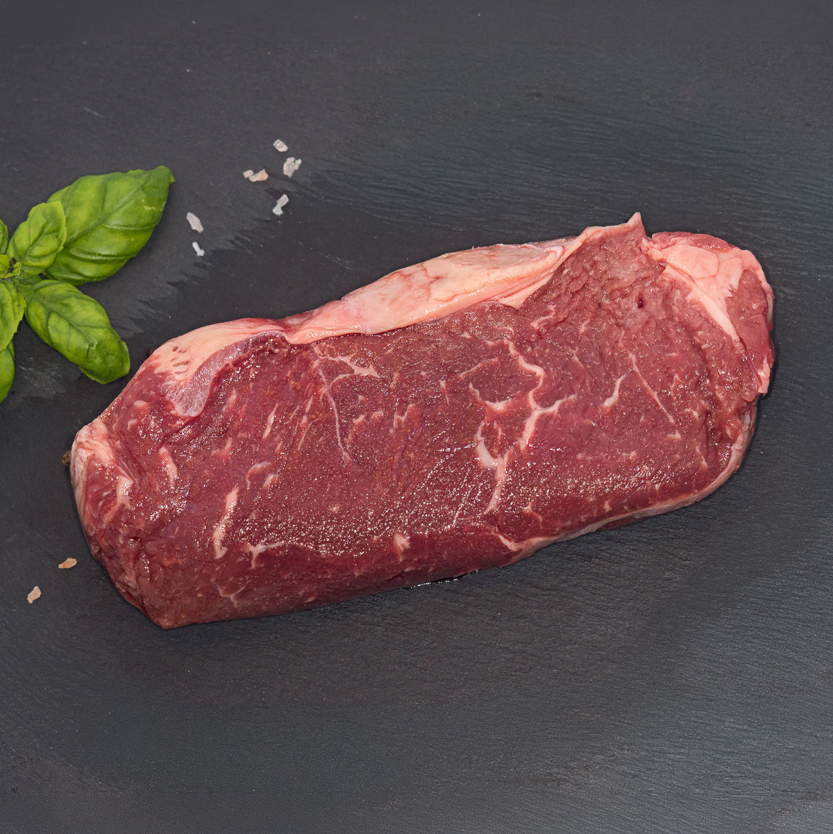 A raw cut of beef steak with marbling, placed on a dark slate surface with a small green herb garnish and a few coarse salt crystals.