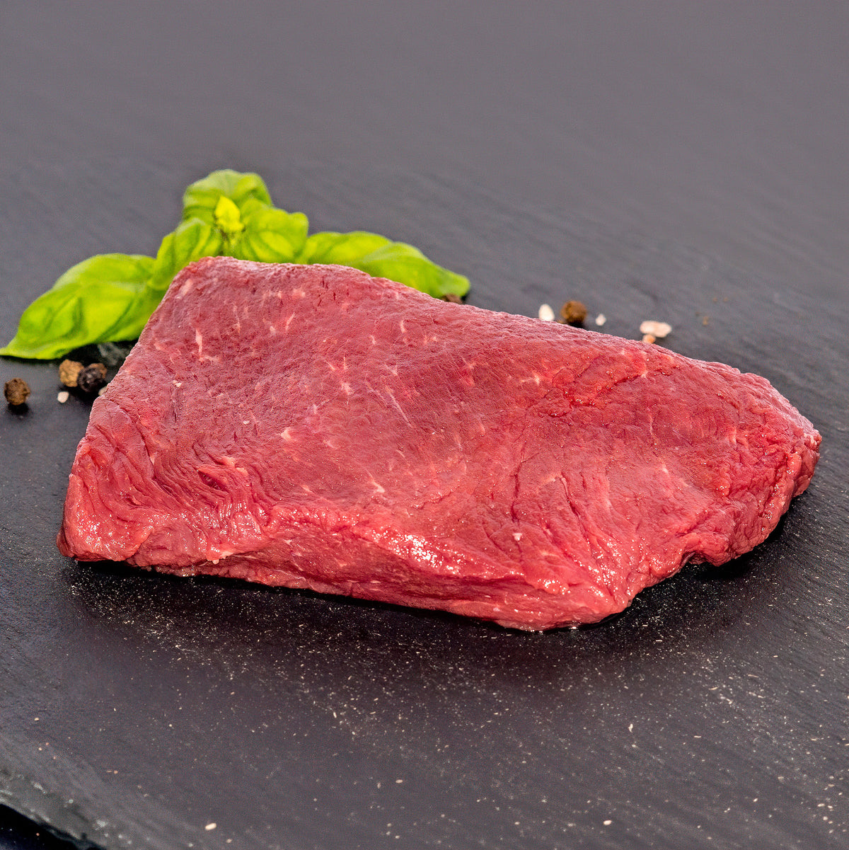 A raw cut of red beef steak on a dark slate plate, garnished with green basil leaves and a few black and white peppercorns.