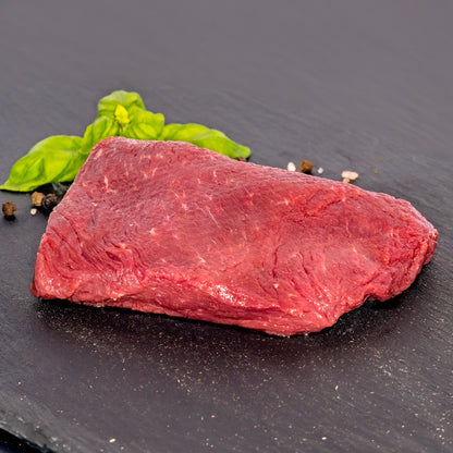 A raw cut of red beef steak on a dark slate plate, garnished with green basil leaves and a few black and white peppercorns.