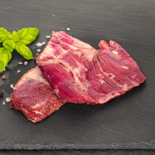 A raw beef skirt steak with visible marbling, arranged on a dark slate surface with a sprig of green basil and scattered coarse salt.