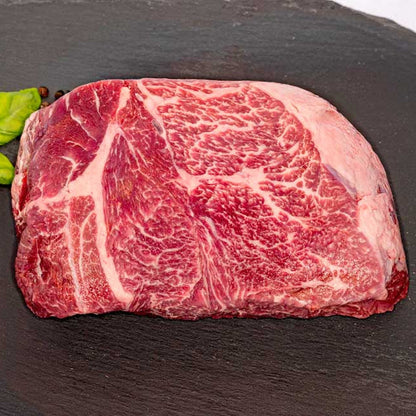 A top view of a well-marbled beef chuck roast, sitting on a black slate oval surface, with fresh basil, peppercorns, and rock salt next to it.