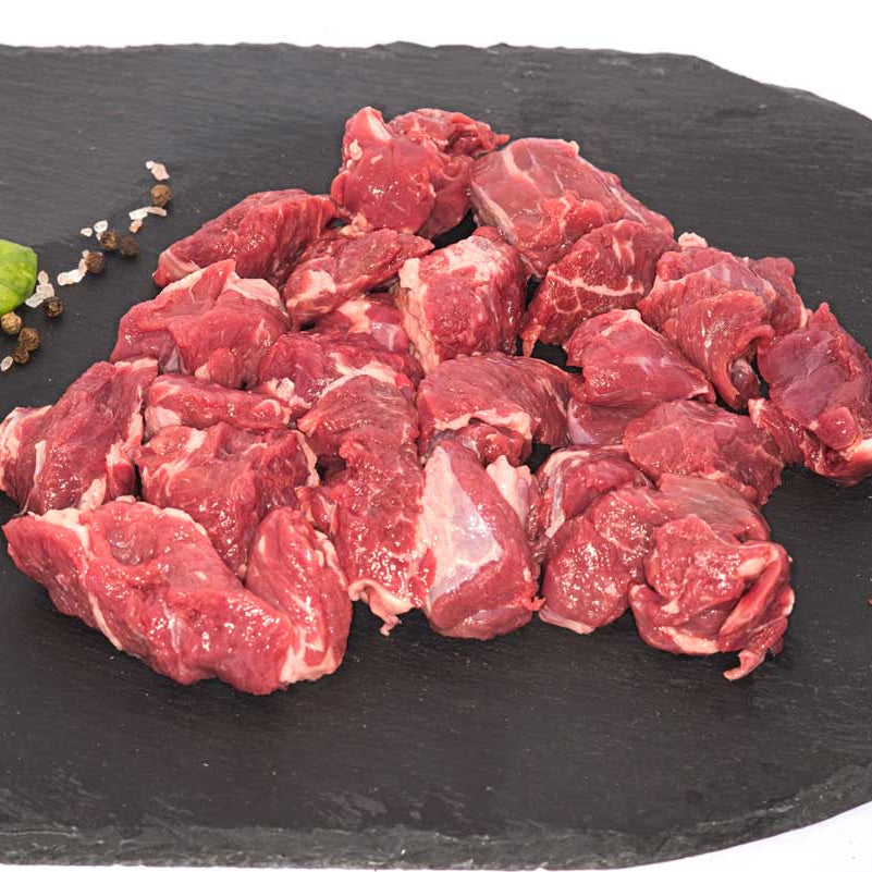 A large pile of cubed beef stew meat, sitting on a black slate surface next to fresh basil, rock salt, and peppercorns.