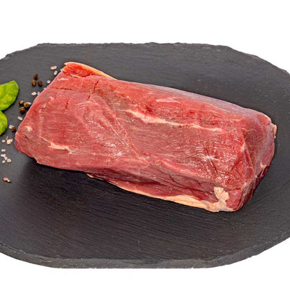 A hefty Eye of Round Beef Roast, sitting on a black slate cutting board, next to fresh herbs, pepper, and rock salt.