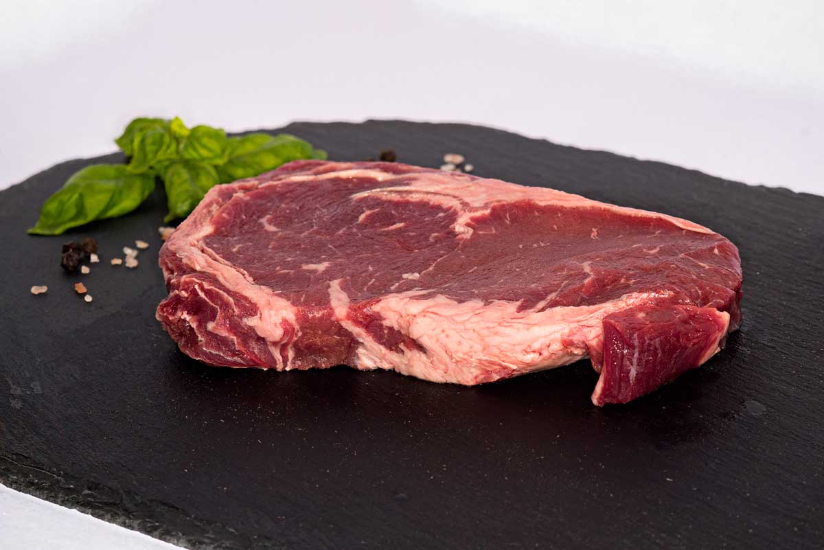 A front view of a boneless beef ribeye steak, sitting on a black slate board with fresh basil, peppercorns, and rock salt in the background.