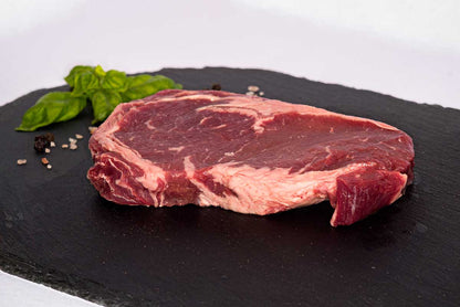 A front view of a boneless beef ribeye steak, sitting on a black slate board with fresh basil, peppercorns, and rock salt in the background.