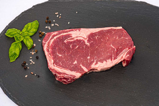 The top view of a boneless beef ribeye steak, sitting on a black slate board with fresh basil, peppercorns, and rock salt next to it.