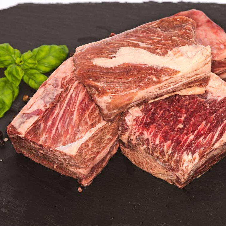 A stack of 4 large cubes of beef short ribs, sitting on a black slate surface next to fresh basil, rock salt, and peppercorns.