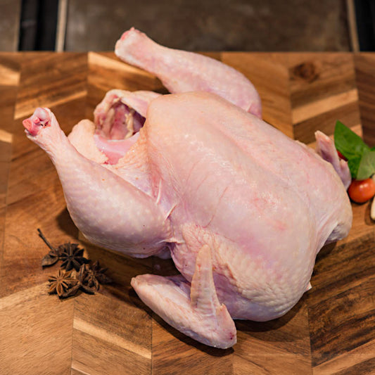 A raw whole chicken laid on a wooden cutting board with star anise and a small tomato and leaf nearby.