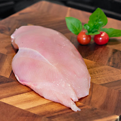 A raw, pale pink chicken breast fillet resting on a wooden cutting board, garnished with fresh green basil leaves and two small red cherry tomatoes.