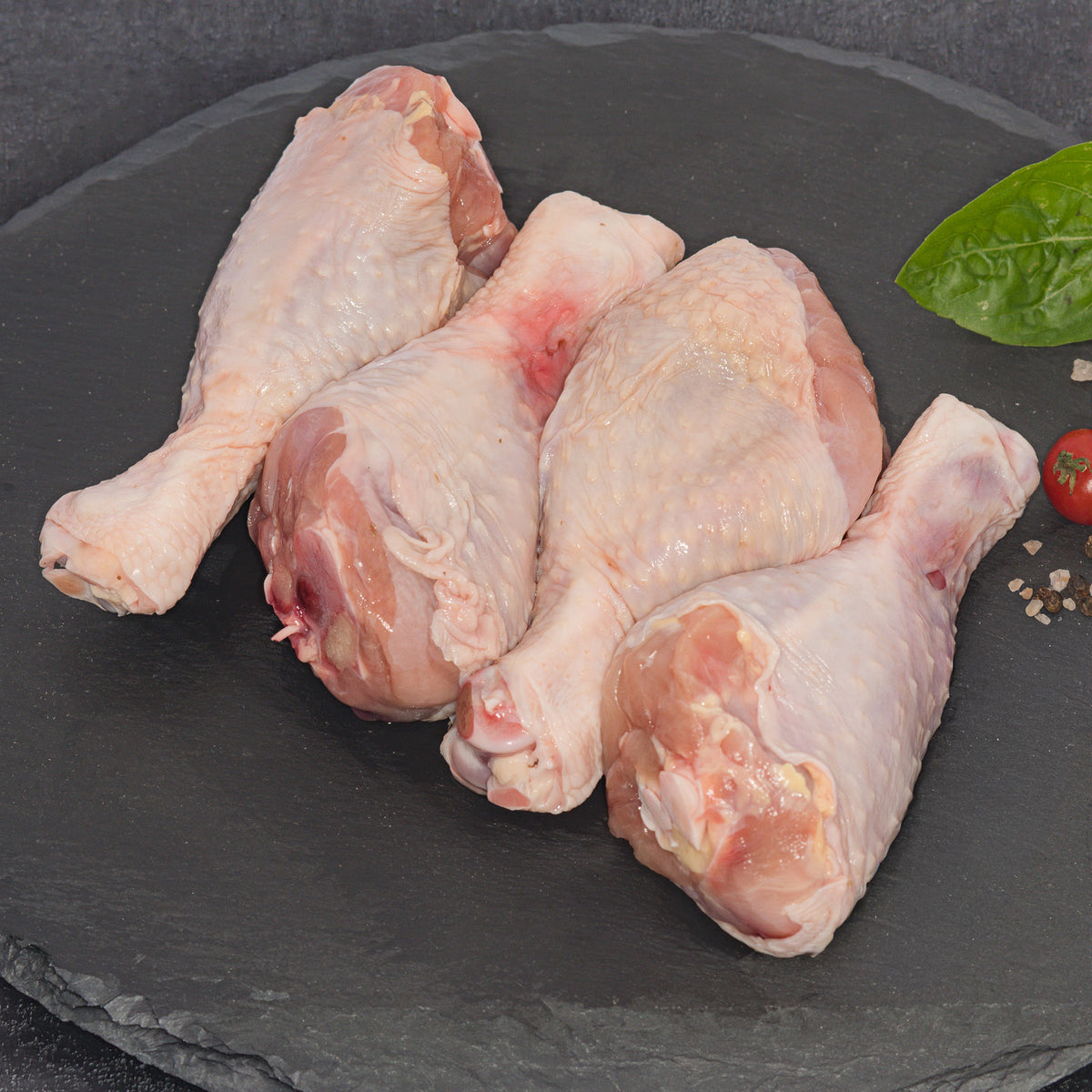 Four raw chicken drumsticks arranged on a dark slate platter with a small green leaf and cherry tomato garnish.